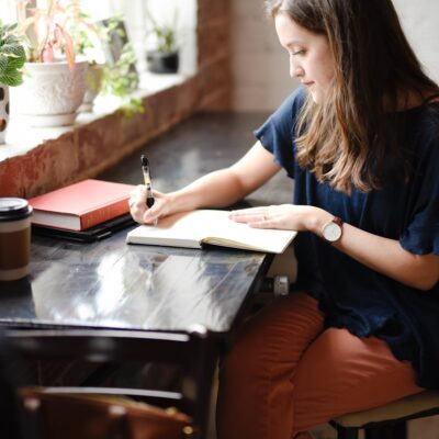 woman sitting in front of black table writing on white book near window