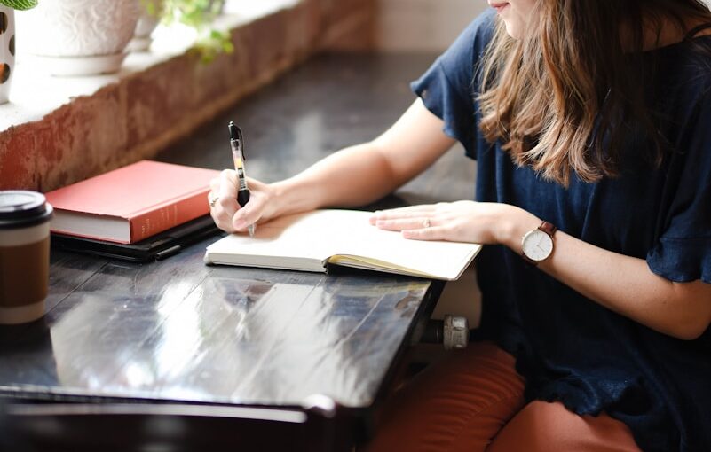 woman sitting in front of black table writing on white book near window